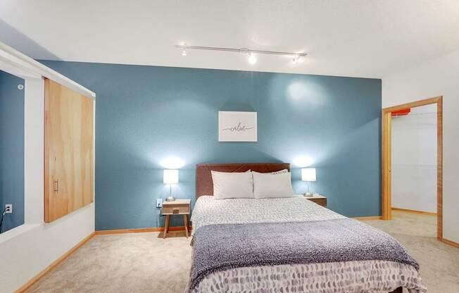 A modern bedroom here at Blue and Lime featuring a bed centered against a bold blue accent wall with neutral bedding and a textured throw. Matching nightstands with table lamps add symmetry, while soft carpeting, track lighting, and warm wood trim create a cozy, well-lit space with an open view into a walk-in closet.