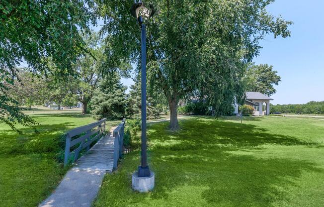 A pathway leading across a small bridge surrounded by lush green grass and trees, with a streetlamp positioned along the path. In the background, a white house is partially visible, adding to the serene landscape under a clear blue sky.