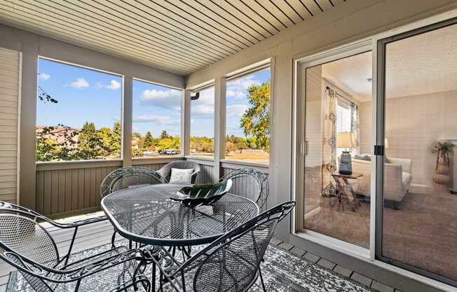A glass table with chairs is on a patio with a view of trees and a house.