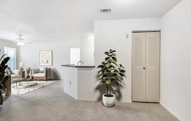 Virtually staged apartment entryway with carpet, indoor plants, livingroom with ceiling fan and light and breakfast bar in the background