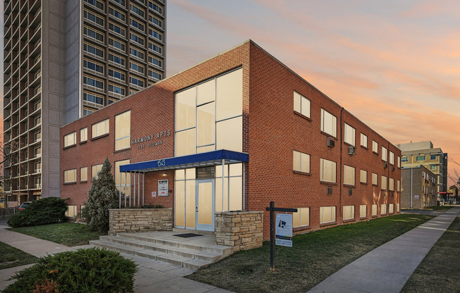 Charming brick apartment building with large windows, surrounded by a well-maintained lawn and urban setting, under a beautiful sunset sky.