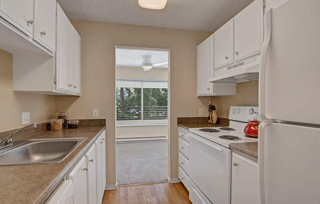 A bright kitchen here at Watercrest with white cabinets, white appliances, and ample counter space, featuring a stainless steel sink and wood-style flooring, opening to a living area with a large window bringing in natural light.