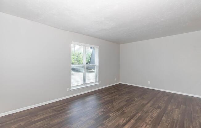 A spacious, empty room with light gray walls and a large window allowing natural light. The floor is made of dark wood, and the overall ambiance is bright and airy, suggesting a fresh, modern living space. The view outside the window shows greenery, indicating a pleasant outdoor setting.
