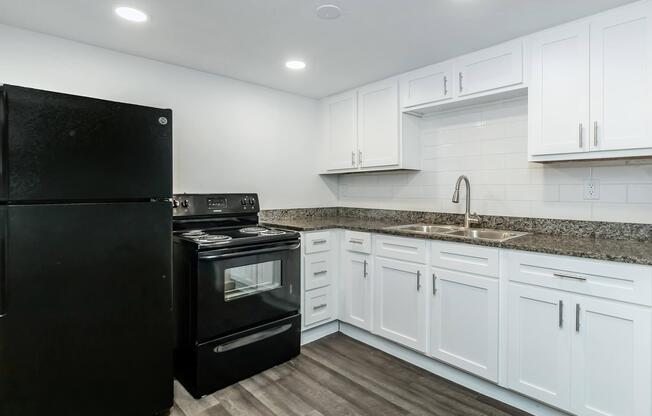 A modern kitchen featuring a black refrigerator and stove, white cabinets, and a granite countertop. The space is well-lit with recessed lighting and has a stainless steel sink with a sleek faucet, set against a white tiled backsplash. The flooring is dark, adding contrast to the overall design.