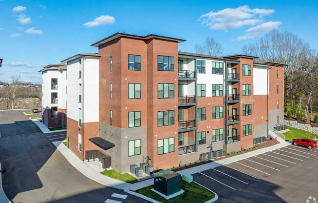 A red car is parked in the parking lot of a multi-story apartment building.