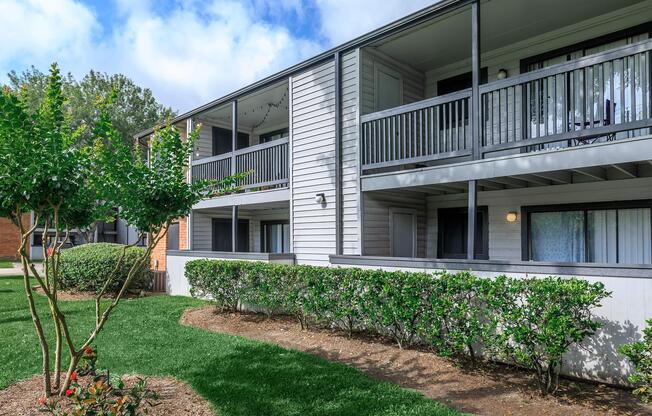 Two-story apartment building with balconies, surrounded by greenery and small trees. The landscape includes well-maintained shrubs and grass, with a partly cloudy sky in the background.