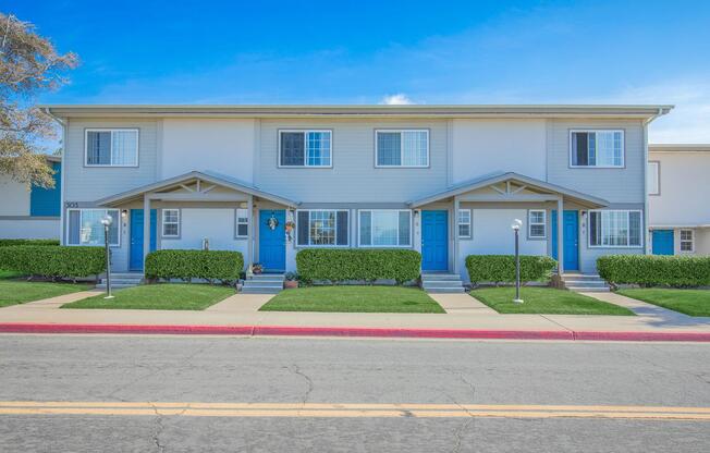 A row of two-story townhouses with blue doors and windows, surrounded by neatly trimmed green bushes and lawn. The sky is clear and blue, and the street in front features a solid dividing line. Pathways lead up to the front doors.