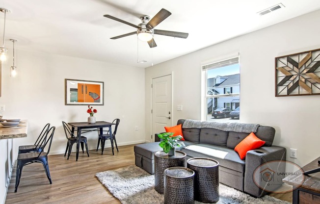 An open window lets light into a white living room with a grey couch, a dining table with chairs, and a ceiling fan.