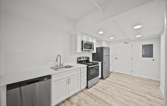 Modern kitchen featuring white cabinetry, stainless steel appliances including a refrigerator, oven, microwave, and dishwasher. The countertops are light-colored, and the flooring is a light wood finish. Bright lighting and a door leading to another area are visible.