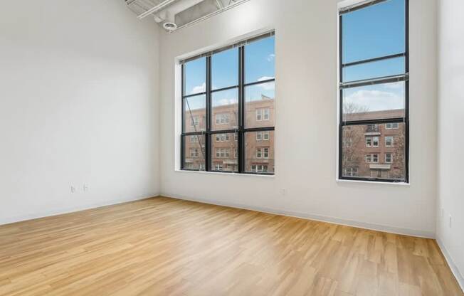 A room with wooden floors and two windows showing a view of a building.