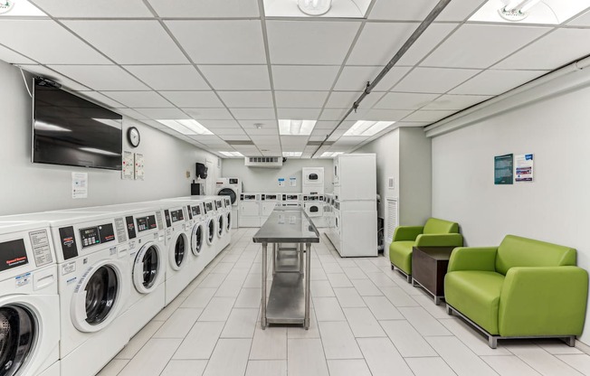 A laundromat with a row of washers and dryers on the left and a table and two chairs on the right.