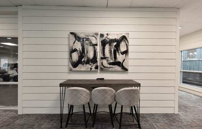 A black and white abstract painting hangs on a white wall above a dark wooden table with four grey chairs.