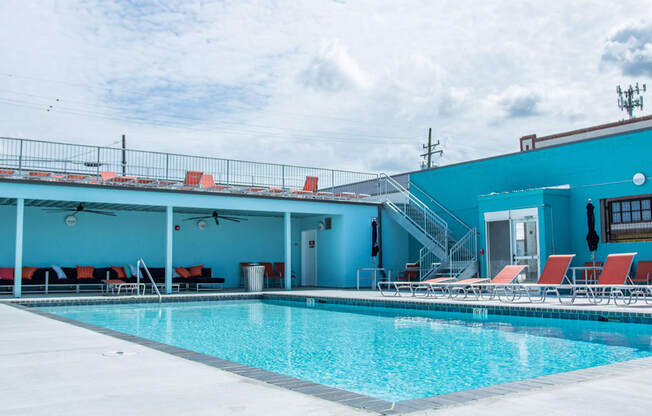 A swimming pool with red lounge chairs and a blue building in the background.