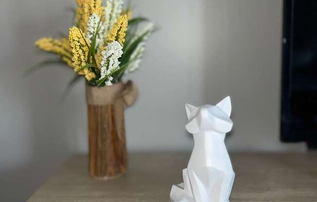 A white ceramic fox figurine sits on a wooden surface next to a vase with yellow flowers.