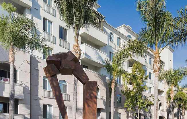 A sculpture of a man holding a woman is in the foreground of a grassy area in front of a building with palm trees. at La Jolla Crossroads Apartments, California