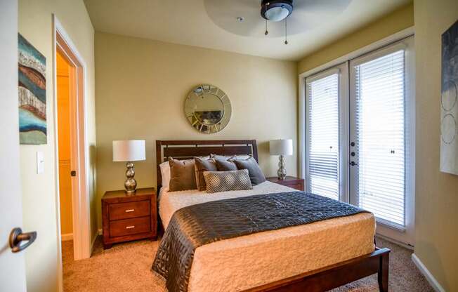 A cozy bedroom here at Andalucia Villas with beige carpet and soft neutral walls, featuring a wooden bed frame with matching nightstands, warm accent lighting, and natural light streaming through glass doors with white blinds, creating a calm and inviting atmosphere.
