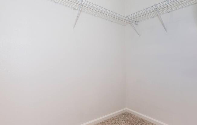 A clean, empty closet with white walls and a beige carpeted floor. Two wire shelves are mounted on the wall, providing storage space. The overall appearance is minimal and organized.