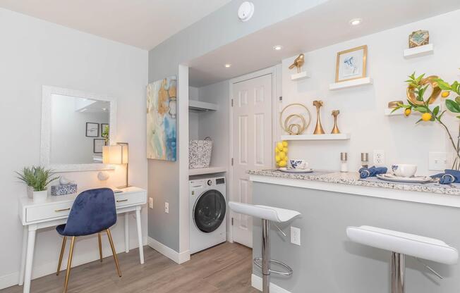 Modern interior featuring a bright and airy space with a sitting area, a mirrored desk with a blue chair, and a laundry area with a washing machine. The decor includes stylish shelves with decorative objects and fresh lemons, and a stylish countertop with bar stools, all set against light-colored walls.