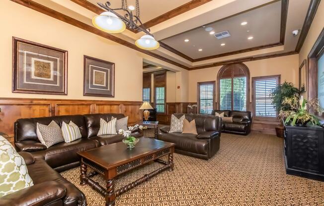 A cozy living room featuring dark leather sofas with decorative pillows, a wooden coffee table, and stylish wall art. The warm interior includes wooden detailing, soft lighting from hanging fixtures, and large windows with plantation shutters. Decorative plants add a touch of greenery to the space.