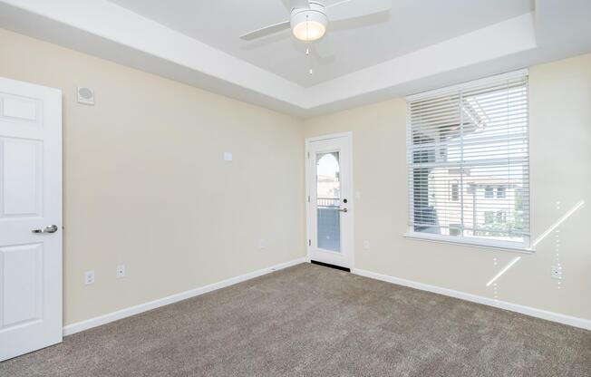 Interior of a vacant room featuring light beige walls, a ceiling fan, and a large window with blinds. The floor is carpeted in a light brown shade, and a door leads outside. The room has a simple and modern design, with no furniture present.