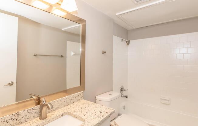 A clean and modern bathroom featuring a bathtub with a white tiled surround, a granite countertop with a sink, a large mirror above the sink, and a towel rack on the wall. The walls are painted in a light gray color, and there is natural light coming from an adjacent room.