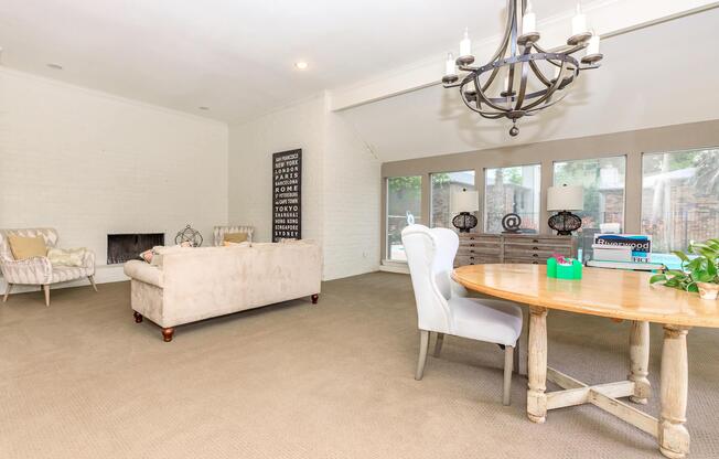 A spacious living room featuring a light-colored sofa, a cozy armchair, a wooden dining table with chairs, and large windows allowing natural light. The decor includes a wall with framed text art and a potted plant on the table.
