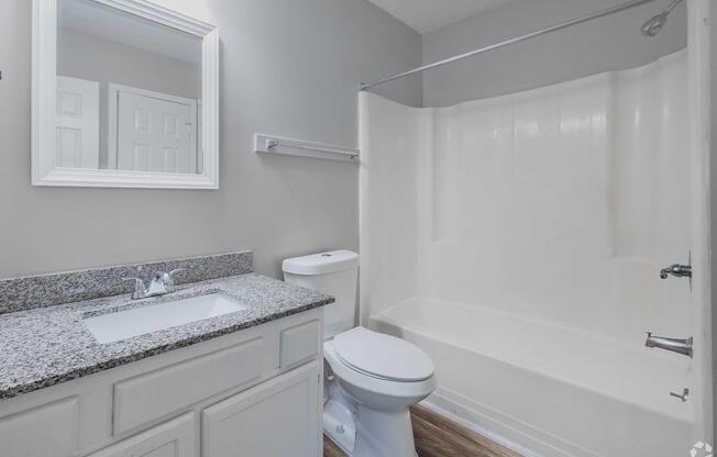 A modern bathroom featuring a white bathtub, a granite countertop sink with a mirror above, and a toilet. The walls are painted in a light gray color, and there is a towel bar mounted on the wall. The flooring appears to be a wood or wood-like material, contributing to a clean and contemporary aesthetic.