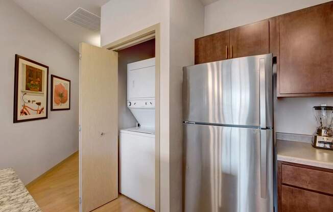 A convenient in-unit laundry area here at Vue at Pinnacle Park with a stacked washer and dryer behind a closet door, next to a stainless steel refrigerator and warm wood kitchen cabinets in a clean, modern layout.
