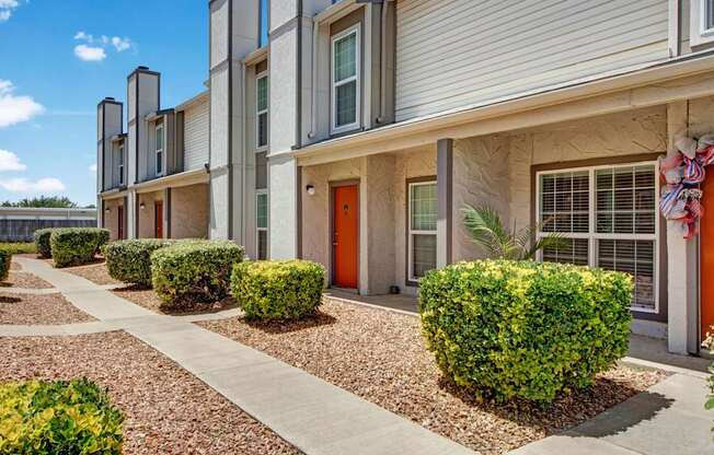 A clean exterior walkway here at High Plains with neatly landscaped shrubs, desert-style gravel beds, and townhouse-style apartment entrances with bright accent doors. The building features large windows and a tidy, welcoming layout under clear blue skies.