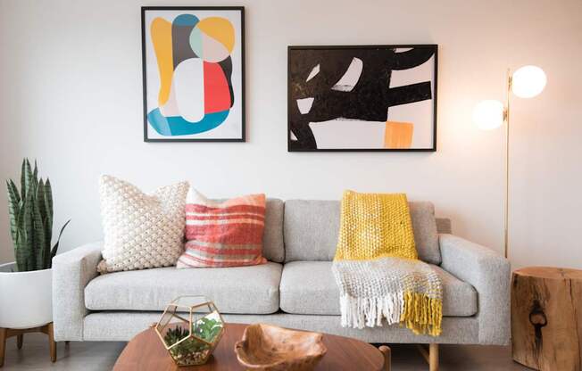 A living room with a grey couch, a yellow throw, and two pieces of abstract art.