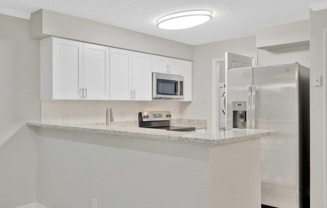 Contemporary kitchen featuring a light-colored granite countertop, white cabinetry, a stainless steel refrigerator, a built-in microwave, and a modern stove. A circular ceiling light illuminates the space, creating a bright and inviting atmosphere.