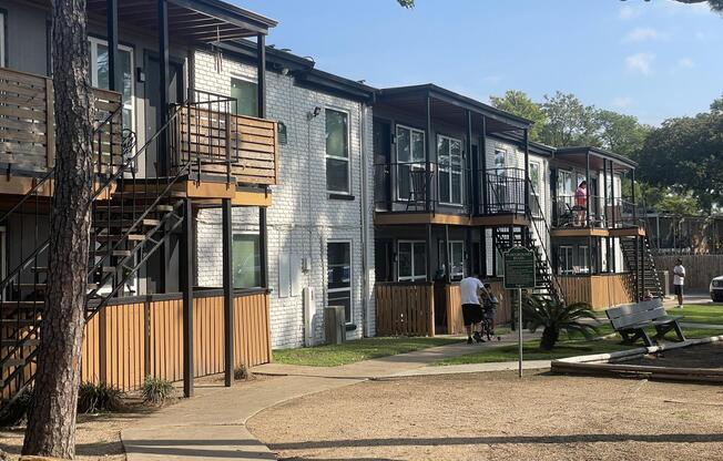 A view of two-story apartment buildings with wooden balconies, surrounded by greenery and a landscaped area. People are seen on the balconies and in the courtyard. A pathway leads through the grass, with trees in the background under a clear blue sky.
