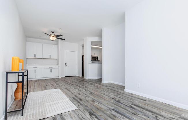 A bright and modern living space featuring light-colored walls and wood-like flooring. A small black console table with decorative items is to the left, and a kitchen area with white cabinets is visible in the background. A ceiling fan hangs above the room, creating a spacious and inviting atmosphere.