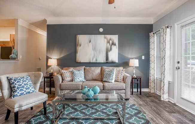 A living room with a grey couch, a blue and white patterned pillow, a glass coffee table, and a painting on the wall.
