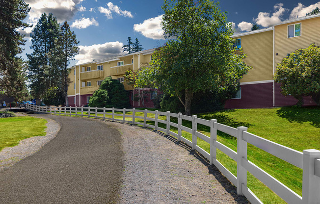 A white fence runs along a gravel road in front of apartment buildings.
