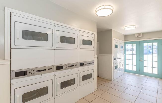 A bright laundry room featuring stacked white washing machines and dryers against a light-colored wall. The space has tiled flooring and is illuminated by overhead lights. There are double doors leading to the outside, with a view of the exit.