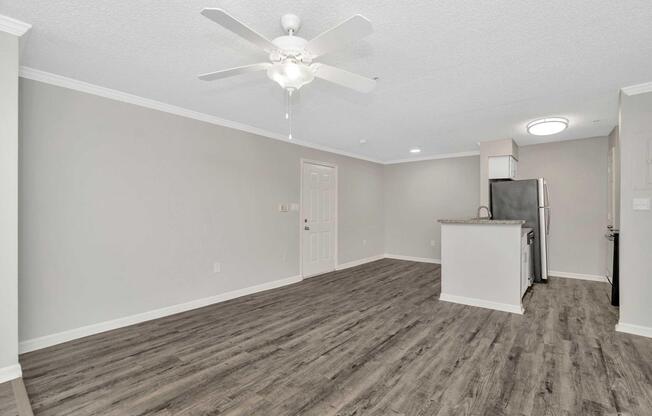 Open living space with light gray walls, featuring laminate flooring. A ceiling fan hangs in the center, and a kitchen area with granite countertop and stainless steel appliances is visible. Natural light illuminates the room, creating a bright and airy atmosphere.