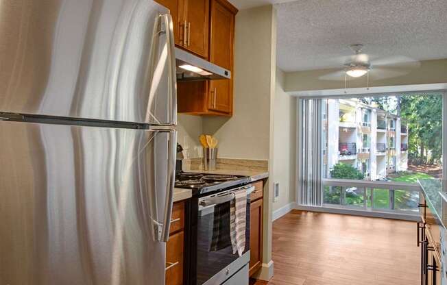 A bright kitchen here at Adagio featuring warm wood cabinetry, light stone countertops, and a full appliance set including a refrigerator, range, and range hood, paired with wood-style flooring and recessed lighting, while an adjacent dining area with a ceiling fan and a large window brings in natural light and views of the surrounding community.