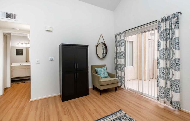 A well-lit interior space featuring a brown cabinet, a green armchair with a decorative pillow, and a large mirror. The room has light wood flooring and a window with patterned curtains. A bathroom area is visible in the background, with two sinks and lights overhead.