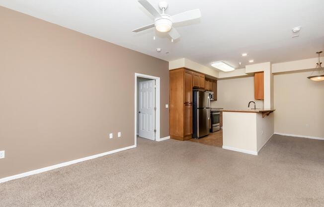 A spacious living area with light brown walls and plush carpet. To the left, a white door leads to another room. In the background, a modern kitchen features wood cabinets, stainless steel appliances, and a countertop with a bar seating area. A ceiling fan is mounted overhead, providing a comfortable atmosphere.