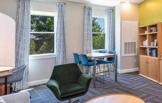The Residences at Crosspoint cyber lounge in Lowell, MA includes a green accent wall with a mounted flat-screen TV, a wooden storage unit with open shelving, a dark green swivel chair, a round wood coffee table, and a high-top table with blue chairs by two curtained windows overlooking trees.