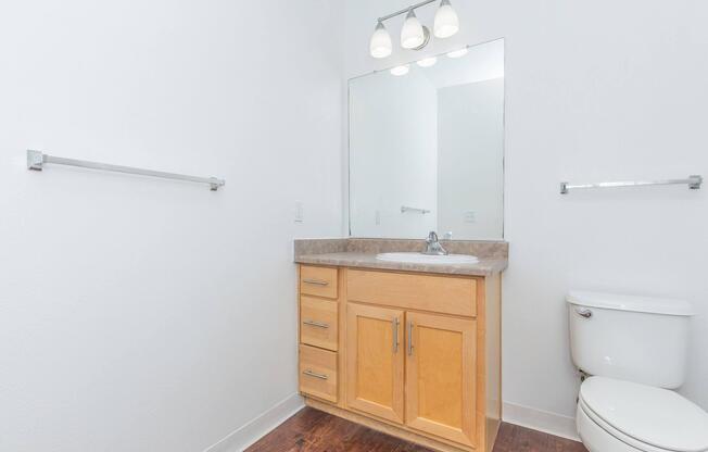 A clean and modern bathroom featuring a light wood vanity with a sink and mirror above, two towel bars on the white walls, a white toilet, and a wooden floor. Soft lighting is provided by three ceiling lights. The space is bright and uncluttered, emphasizing a minimalist design.