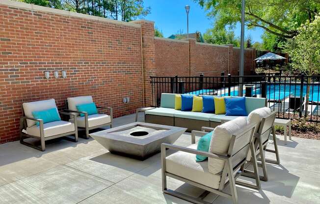 An outdoor community lounge with 4 comfortable chairs, a sofa, and a fire pit near a gated swimming pool.