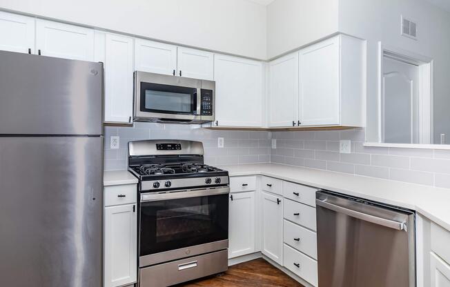 Modern kitchen featuring stainless steel appliances, including a refrigerator, microwave, and gas stove. The cabinets are white, with a sleek countertop and backsplash of light tiles. The floor is a warm wood finish, creating a bright and inviting space.