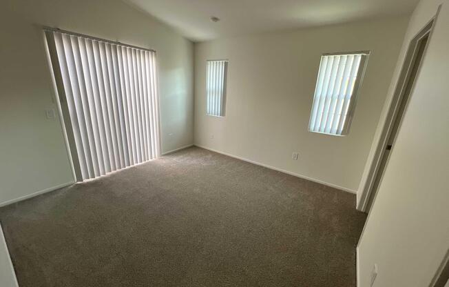 Empty room with light beige carpet and white walls. Two windows with vertical blinds allow natural light to flood the space, creating a bright atmosphere. The room has a simple design with no furniture, making it suitable for various purposes like an office or a living area.