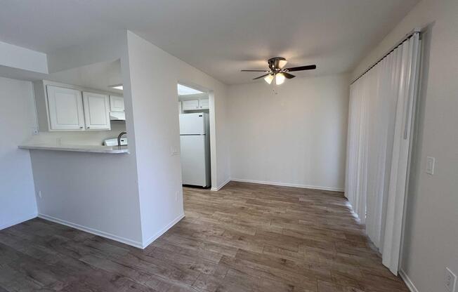 A bright, empty room featuring light brown wood-like flooring, a ceiling fan, and white walls. To the left, there is a kitchen area with a bar counter and a white refrigerator visible. Sheer curtains are positioned at a sliding door that leads outside. The overall atmosphere is clean and spacious.