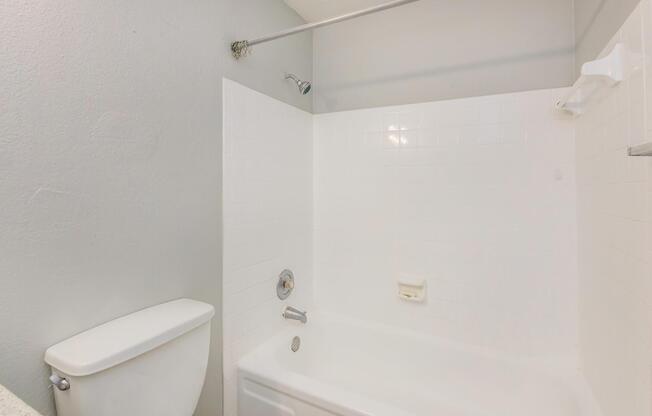 A clean and simple bathroom featuring a white bathtub, showerhead, and a toilet. The walls are painted in a light gray color, and the tiled area around the bathtub is also white, creating a bright and minimalist aesthetic.