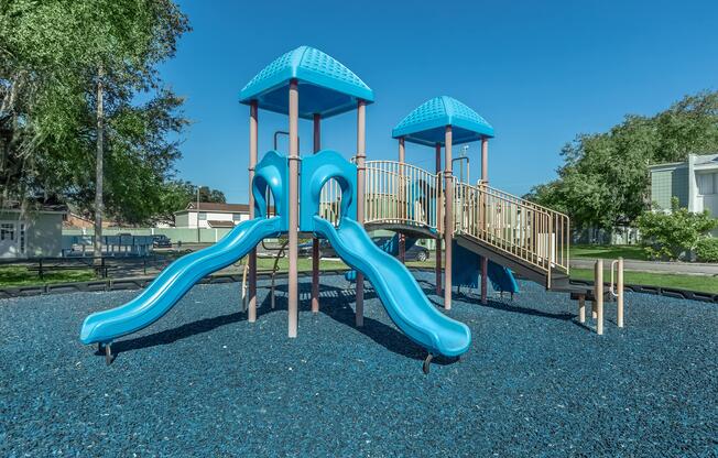 A bright blue playground structure with two slides and a bridge, set on a bed of dark blue rubber mulch. Sunlit trees are visible in the background under a clear blue sky. The playground is designed for children to play and explore.