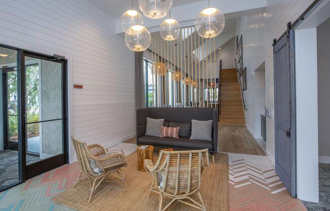 A modern lobby area featuring a cozy sitting arrangement with two woven chairs, a dark gray sofa with decorative pillows, a wooden coffee table, and a large rug. The walls are white shiplap, and large glass doors lead outside. Decorative hanging lights and a staircase are in the background.