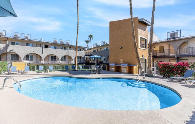 A serene outdoor swimming pool surrounded by palm trees and lounge chairs. The pool features a curved shape, with clear blue water reflecting the sunny sky. Nearby, there are a few shaded seating areas and a building with multiple levels in the background, adding a cozy atmosphere to the scene.